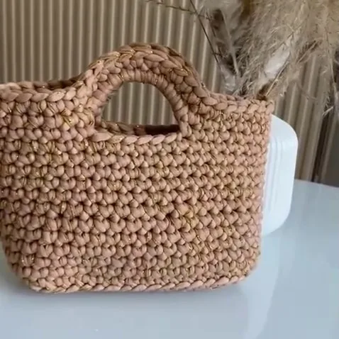 Cabas en crochet beige parcouru d'un fil doré, posé sur une table blanche devant des herbes de la pampa
