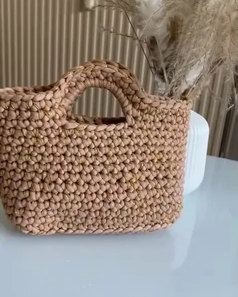 Cabas en crochet beige parcouru d'un fil doré, posé sur une table blanche devant des herbes de la pampa