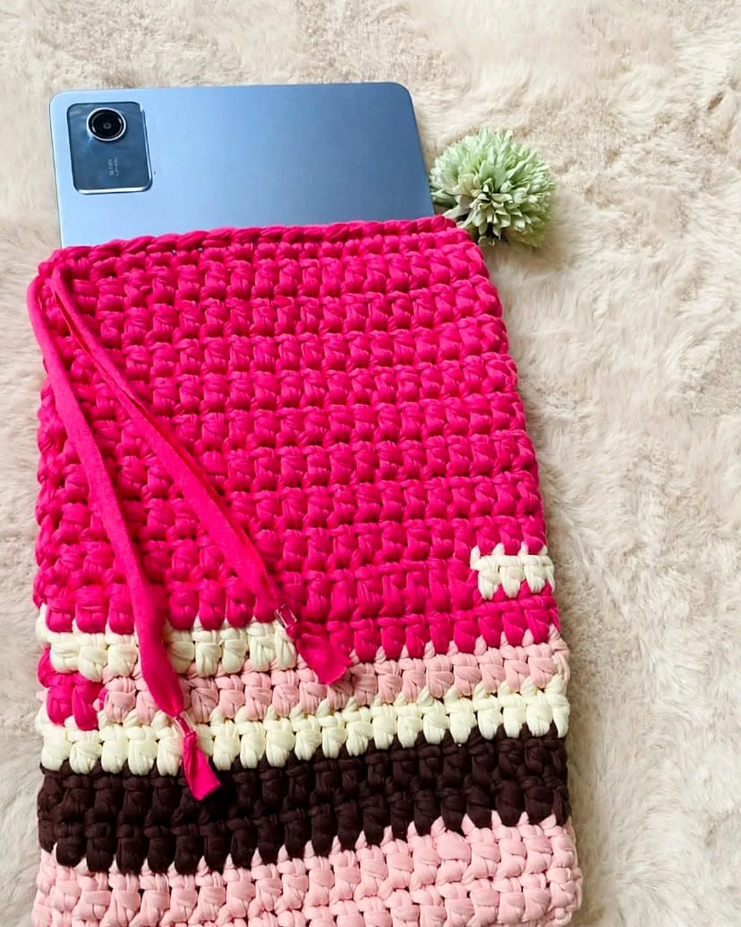 Pochette en crochet rose fuchsia rayée, une tablette glissée à l'intérieur, posée sur un tapis crème